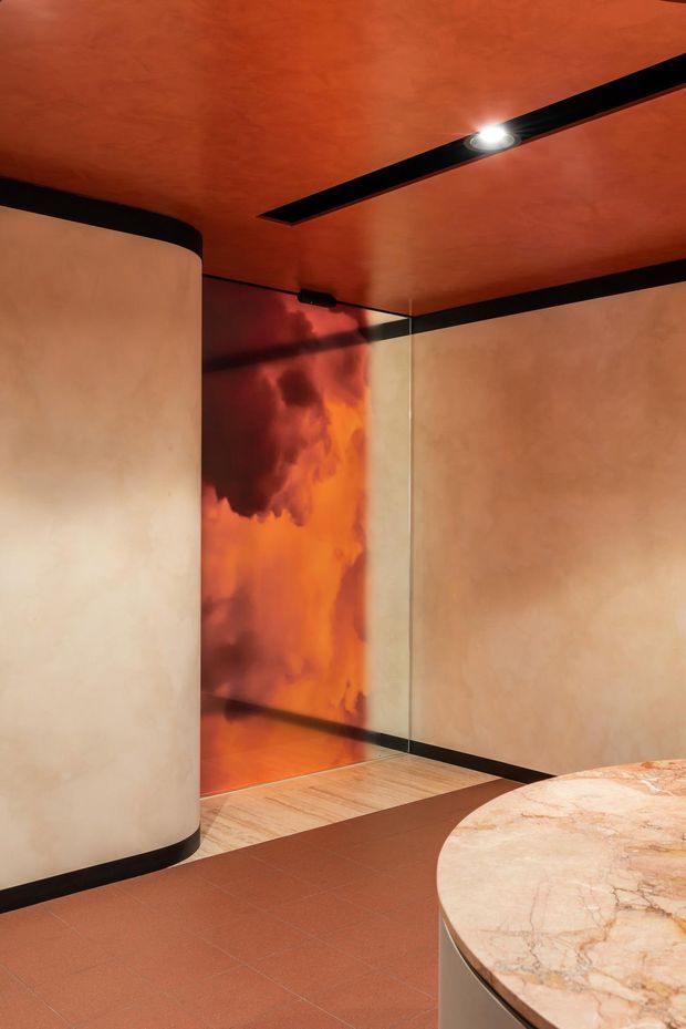 A dramatic, highly stylised interior corner with curved pale plaster walls, a terracotta-orange ceiling and tiled floor, black linear trims and track lighting, and a reflective orange-toned feature wall or glazed opening. A rounded marble-topped built-in counter or table occupies the foreground. The space has a warm, moody, gallery-like atmosphere with strong geometric detailing.