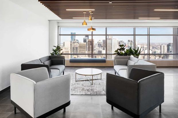A bright contemporary high-rise lounge or reception area with pairs of grey upholstered armchairs arranged around a round coffee table and patterned rug. Full-height windows provide expansive city views, while a dark linear timber slat ceiling, polished light floor, indoor plants, and warm brass pendant lights add contrast and refinement.