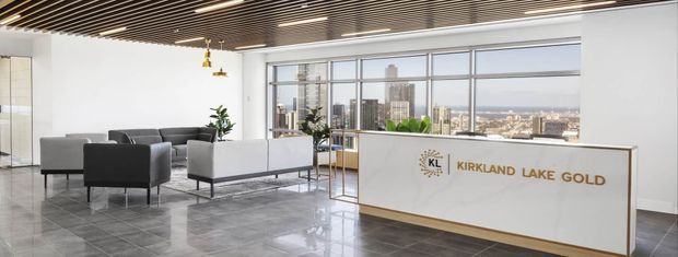 A bright, polished commercial reception lounge with grey armchairs and a white built-in reception desk bearing the “Kirkland Lake Gold” logo. Floor-to-ceiling glazing provides city views, while a dark linear slatted ceiling, recessed strip lighting, reflective stone-look flooring, and sparse greenery create a contemporary corporate interior.
