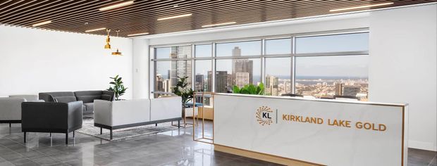A bright contemporary commercial reception and lounge area with floor-to-ceiling windows overlooking a city skyline. The space features a white reception desk with gold trim and the text “Kirkland Lake Gold,” grey lounge seating, polished grey flooring, a timber slatted ceiling, and modern pendant lighting.