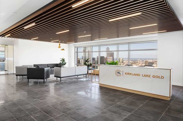 A bright, modern commercial reception lobby with a white reception desk bearing the Kirkland Lake Gold logo, grey lounge seating, large city-facing windows, polished dark tiled flooring, and a striking suspended timber slat ceiling with integrated linear lighting.