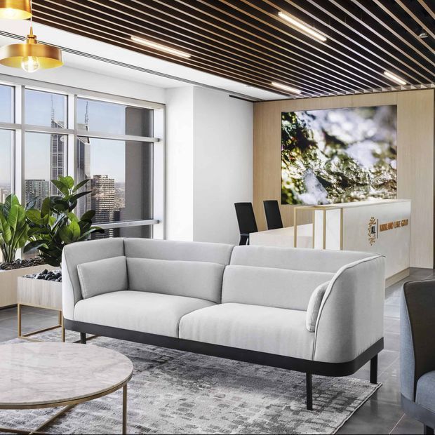 A bright contemporary commercial reception lounge with a large light-grey upholstered sofa, marble-topped round coffee table, patterned rug, indoor planting, and a reception counter. Floor-to-ceiling windows provide city views, while a timber slatted ceiling with integrated linear lighting and a large digital display create a refined corporate interior.