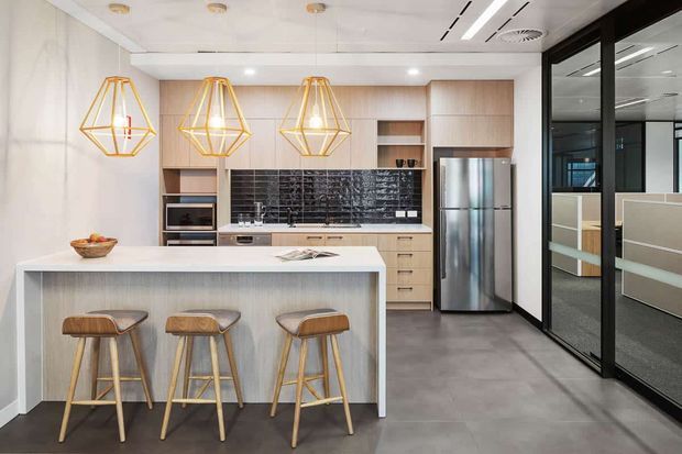 Contemporary open-plan kitchen with a large white island and three timber bar stools, pale timber cabinetry, dark glossy tile splashback, stainless-steel appliances, and geometric brass pendant lights. Black-framed glazed partitions on the right reveal an adjacent office or meeting area.