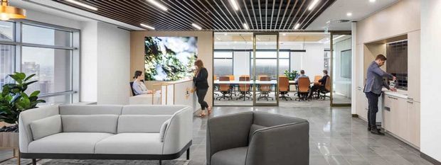 A contemporary commercial office interior with a lounge and reception area in the foreground, featuring grey upholstered sofas, polished light-grey tiled flooring, white walls, extensive glazing, and a timber-look slatted ceiling with linear lighting. A meeting room and open-plan workstations are visible beyond the lounge, along with built-in cabinetry and a refreshment area.