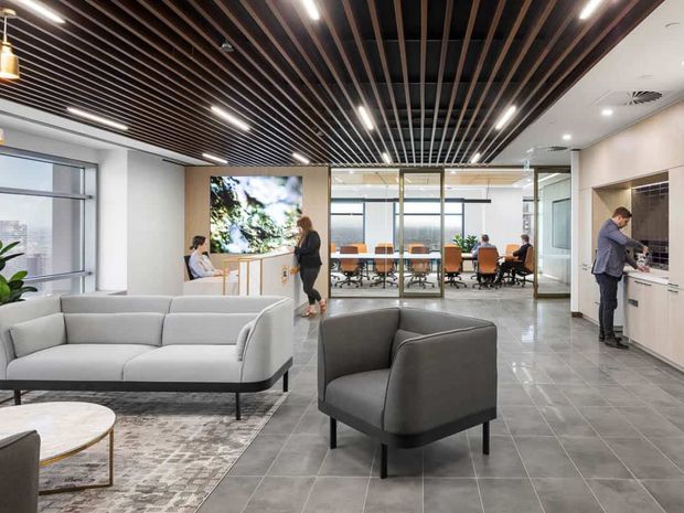 Contemporary open-plan commercial office interior with a lounge reception area in the foreground, a glass-enclosed meeting room beyond, and a compact kitchenette or refreshment zone to the right. The space features a dark timber-look slatted ceiling with integrated linear lighting, pale walls, large windows, grey tiled flooring, and soft grey upholstered seating.