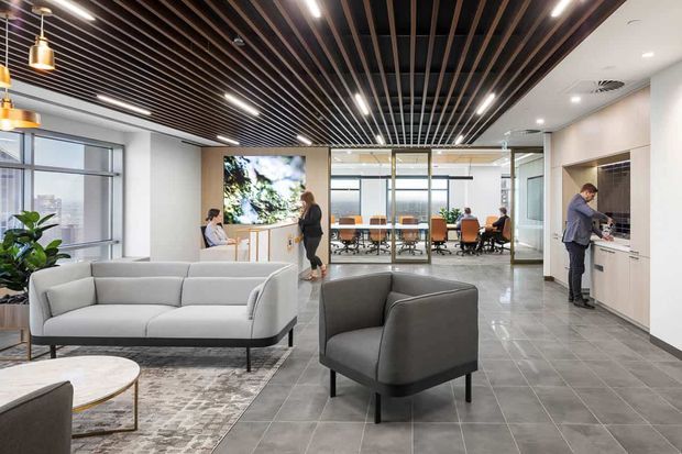 A contemporary commercial office interior with a reception or lounge area in the foreground, furnished with light grey sofas and armchairs over a patterned rug. The space has large grey tiled flooring, white walls, extensive glazing, a timber slatted ceiling with integrated linear lighting, pendant lights, and a glass-enclosed meeting room in the background. A kitchenette or refreshment area is visible on the right.