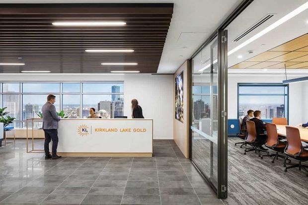 A bright contemporary commercial office reception area with a white reception desk bearing the Kirkland Lake Gold logo, dark wood slatted ceiling panels, large windows overlooking a city skyline, grey tiled flooring, and a glazed meeting room with conference seating visible to the right.