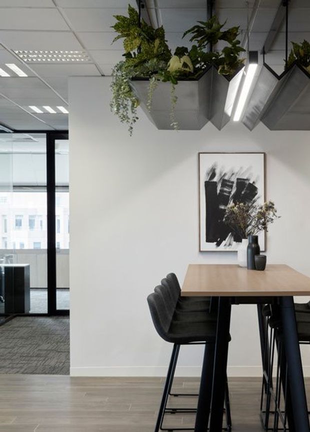 A contemporary office dining or breakout area with a long light-wood communal table and black high-back chairs. White walls, timber-look flooring, exposed ceiling tiles, black-framed glass partitions, geometric suspended planter boxes, linear LED lighting, and monochrome artwork create a refined modern workplace interior.
