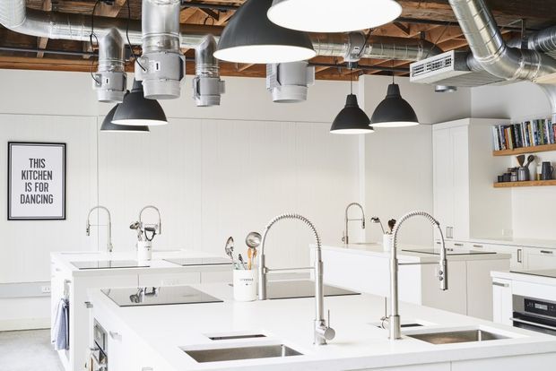 Bright contemporary commercial-style kitchen with multiple large white islands, integrated sinks and tall gooseneck faucets. The room has white cabinetry and wall panelling, exposed timber ceiling structure, extensive exposed metal ductwork, and multiple black-and-white pendant lights.