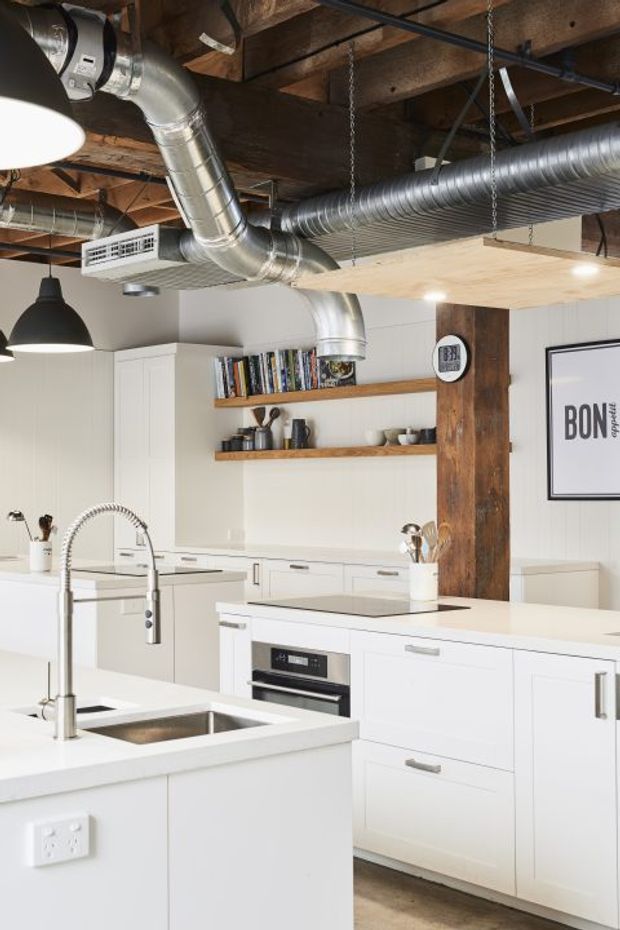 Bright contemporary kitchen with extensive white cabinetry, multiple islands, stainless-steel sinks and pull-out mixer taps, integrated oven, timber open shelving, and exposed timber beams. Large exposed metal ventilation ducts and industrial pendant lighting create a strong industrial loft character, while a substantial timber post and warm timber flooring add contrast.