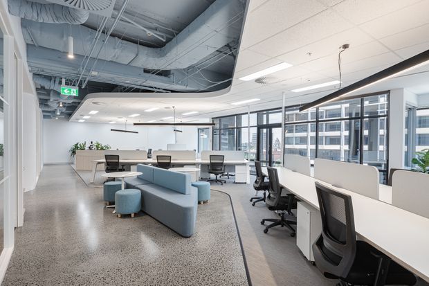 Bright contemporary open-plan commercial office with rows of workstations, ergonomic office chairs, curved acoustic screens, informal blue upholstered seating, extensive glazing, polished concrete flooring, and an exposed services ceiling in part of the space.