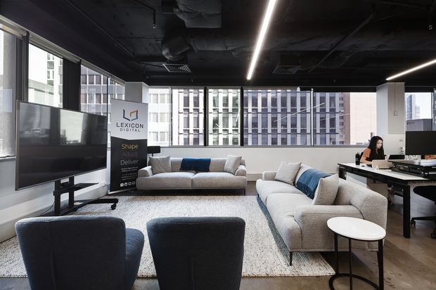 A contemporary open-plan office lounge and meeting area with large windows overlooking neighboring urban buildings. The space features a black exposed ceiling with linear LED lighting, pale carpet, grey and blue sofas, dark lounge chairs, a round side table, and workstations with monitors. A Lexicon Digital sign and employee workspace are visible in the background.