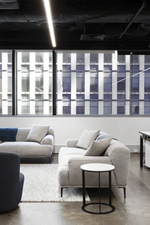 A bright contemporary lounge seating area with two light grey sofas and a dark accent armchair arranged around a small round side table. The room has polished concrete flooring, a large pale rug, expansive black-framed windows, and an exposed black ceiling with linear lighting and visible services. A neighbouring modern office building is visible through the glazing.