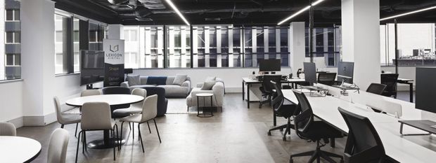 Open-plan contemporary office interior with long white workstations, multiple ergonomic office chairs, computer monitors, meeting tables, and a lounge seating area. The space has exposed dark services at the ceiling, linear strip lighting, white structural columns, large windows, and a polished light-toned floor.