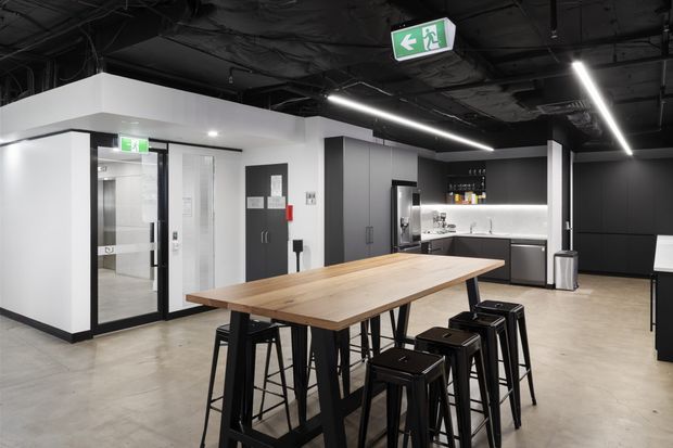 Modern commercial-style breakout or staff kitchen area with a long timber communal table and black bar stools, polished concrete flooring, black exposed ceiling services, linear LED lighting, dark cabinetry, and a compact kitchenette at the rear. A glazed entry or lift lobby is visible on the left, with emergency exit signage overhead.