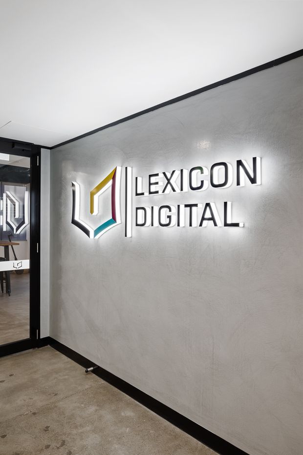 A modern commercial interior entry area featuring a backlit “LEXICON DIGITAL” logo mounted on a textured light-grey wall. Black trim, a black-framed glazed doorway, and a polished concrete floor create a clean, contemporary office or studio reception aesthetic.