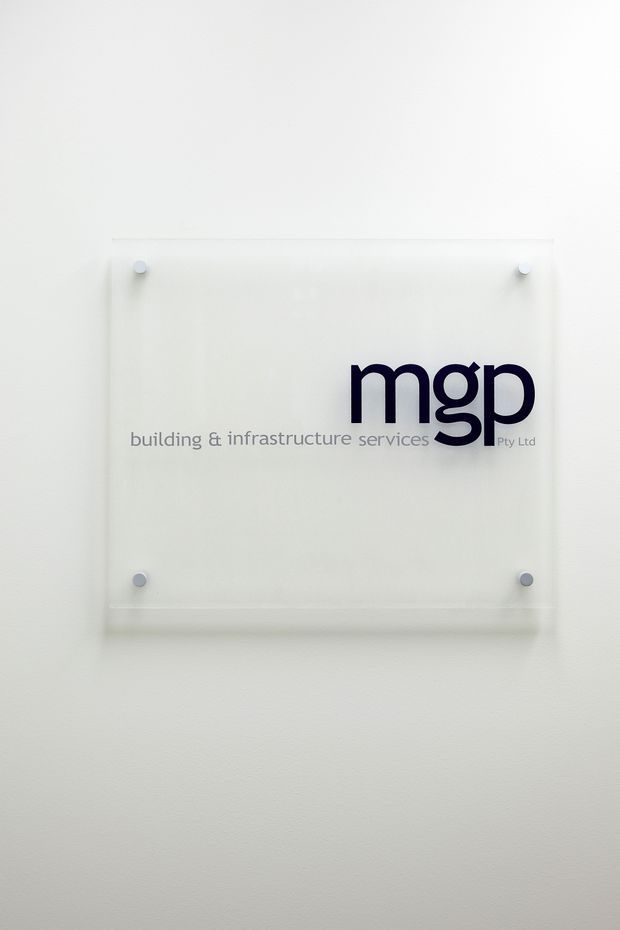 A minimalist corporate wall sign mounted on a white painted wall. The sign is a clear acrylic panel secured with four round metal or silver fixings and displays the logo and text “mgp building & infrastructure services Pty Ltd” in dark blue and grey lettering.