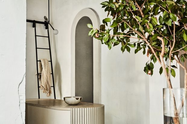 A refined, softly lit interior vignette featuring a curved pale plaster wall niche, rounded ribbed built-in cabinetry or console, and a tall black metal ladder-style towel rack with a small pendant light. A large leafy indoor tree in a clear vessel adds an organic element, while neutral finishes and sculptural forms create a calm contemporary atmosphere.