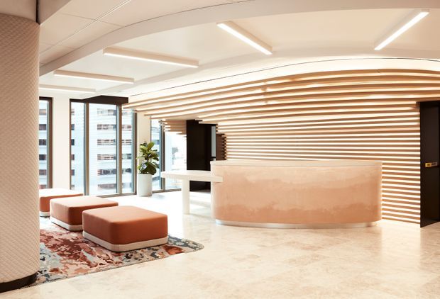 A bright contemporary reception lobby with a curved pale stone reception desk, a dramatic horizontal timber slat feature wall and ceiling, polished light flooring, linear LED lighting, floor-to-ceiling glazing, and rounded rust-coloured lounge ottomans on a patterned rug. The space has a refined, welcoming commercial interior with a sculptural, softly curved layout.