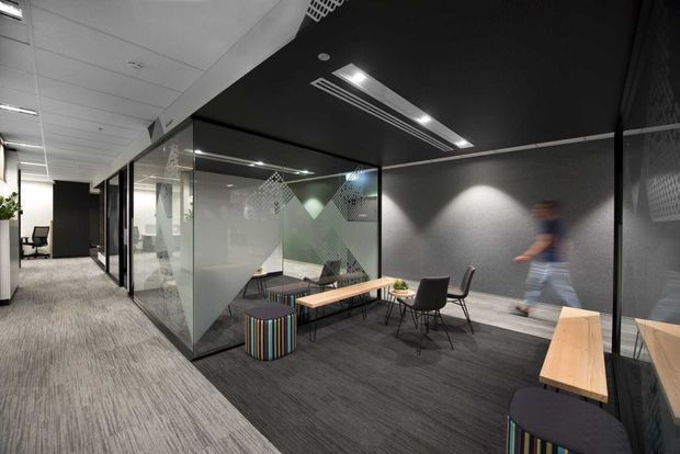 Contemporary commercial office interior showing a carpeted corridor beside a glazed meeting or breakout room. The space features black acoustic ceiling panels, recessed linear lighting, dark grey walls, patterned privacy film on glass partitions, timber tables, dark chairs, and colourful striped stools. A blurred person is walking through the space.