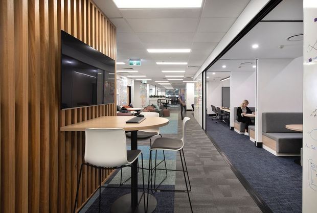 A contemporary open-plan commercial office interior with a long circulation aisle, meeting rooms enclosed by full-height glass partitions, shared workstations, booth seating, and a small collaboration area. A timber vertical-slat feature wall supports a wall-mounted television beside a round communal table and stools. The space has carpet flooring, suspended acoustic ceiling tiles, recessed linear lighting, and a clean professional fit-out.