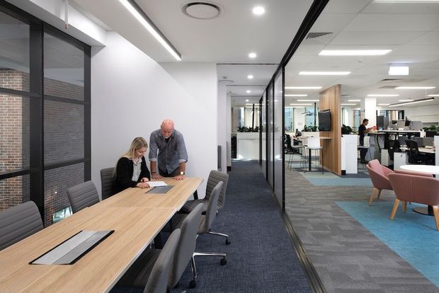 A bright contemporary commercial office interior featuring a long timber meeting table with upholstered office chairs in the foreground, separated from an open-plan workspace by a black-framed glazed partition. The office includes carpeted flooring, white walls and ceilings, linear LED lighting, recessed downlights, exposed services, built-in timber joinery, and informal lounge seating in the background.