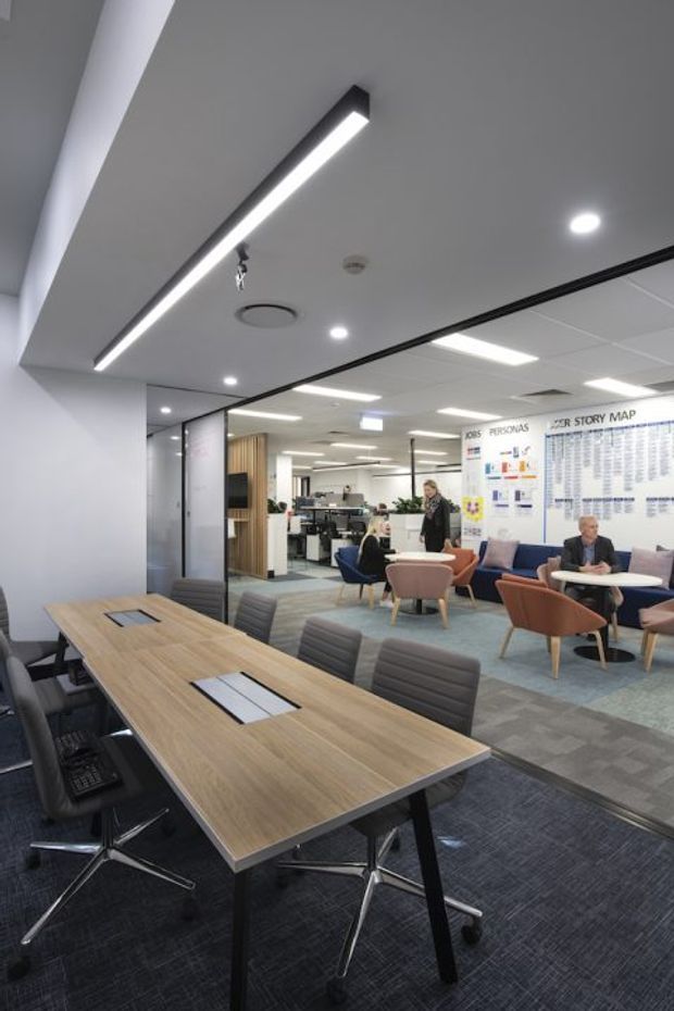 A contemporary open-plan commercial office interior with a large timber meeting table and black rolling chairs in the foreground. The space includes carpeted flooring, suspended linear and recessed lighting, glazed partitions with black frames, a reception or collaboration area, lounge seating, workstations, and a large wall-mounted organisational graphic.