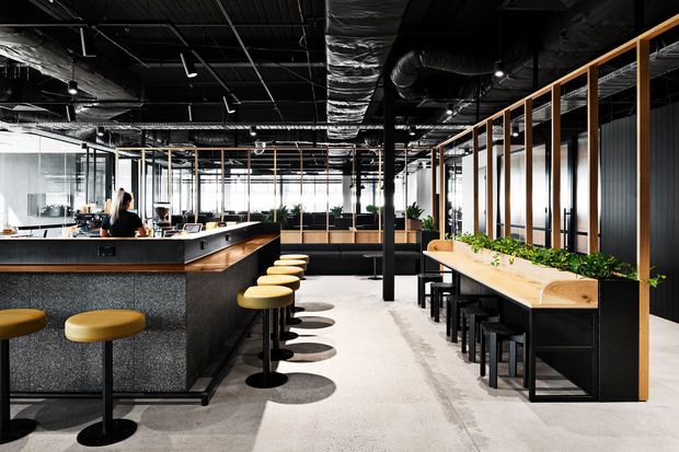 A contemporary open-plan commercial interior resembling a café, hospitality venue, or workplace amenity area. The space features a large dark island counter with mustard-yellow stools, a long timber communal counter with integrated planter, black stools, exposed black ceiling services and ductwork, glass partitions, and polished concrete flooring. The overall scheme is industrial and modern with strong black, timber, and yellow accents.