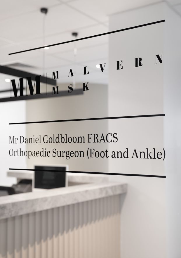 Minimalist medical practice reception area viewed through a glass partition. Black typography on the glazing identifies Malvern MSK and an orthopaedic surgeon, while a pale stone reception counter with vertically ribbed cabinetry sits behind it. The space has white walls, a simple suspended ceiling, black linear detailing and understated contemporary finishes.