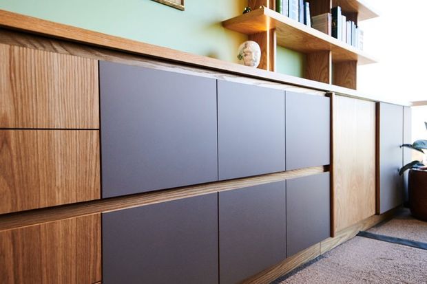 Close-up view of a built-in timber storage and shelving unit, with warm wood-grain cabinetry, matte dark grey drawer and door fronts, open display shelves with books, and a soft green wall behind. A potted plant and partial rug are visible at the edge of the room.