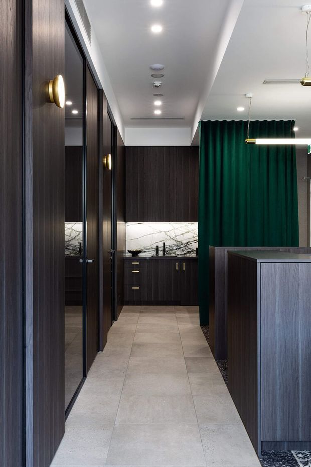 A refined commercial interior hallway or bathroom vanity area with dark vertically grained timber-look wall panelling and cabinetry, full-height mirrored doors on the left, a pale large-format tiled floor, and a dramatic white marble-look splashback at the rear vanity. Deep emerald green curtains and warm brass wall sconces provide contrast, while recessed ceiling lights and linear pendant lighting create an upscale, contemporary atmosphere.