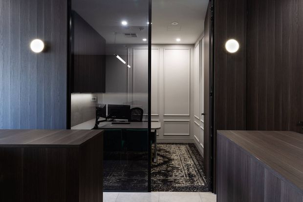 A compact, sophisticated home office viewed through a glazed opening, with dark wood wall panelling and built-in cabinetry framing the space. The office contains a desk, task lighting, shelving or storage, and a patterned rug, while the adjoining passage features white decorative wall panelling and concealed doors.