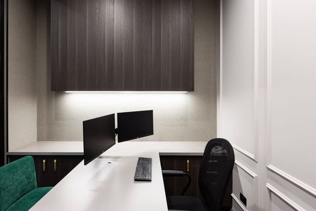 Compact, refined home office with a white built-in desk, dual black monitors, ergonomic office chair, and a green upholstered chair. Dark wood overhead cabinetry with integrated under-cabinet lighting contrasts with light textured walls and white decorative wall panelling.