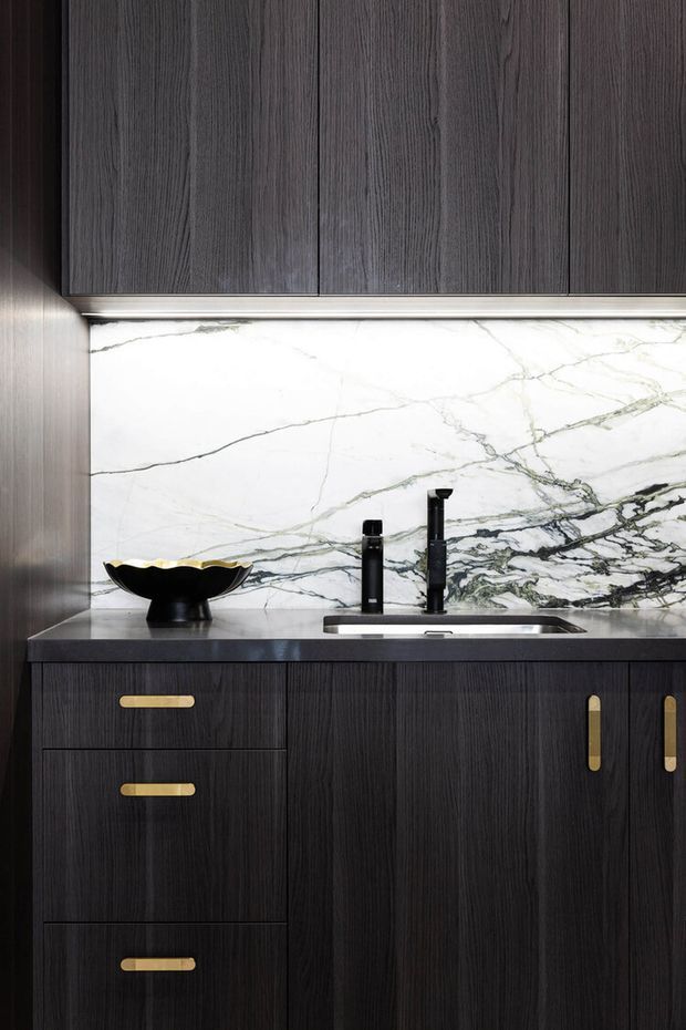 Close-up of a refined kitchen or kitchenette joinery installation featuring dark vertically grained timber cabinetry with slim brass-gold handles, a dark benchtop, and a dramatic white marble backsplash with grey-green veining. A black undermount sink and matte black tapware sit beneath concealed under-cabinet lighting. A decorative black bowl rests on the benchtop.