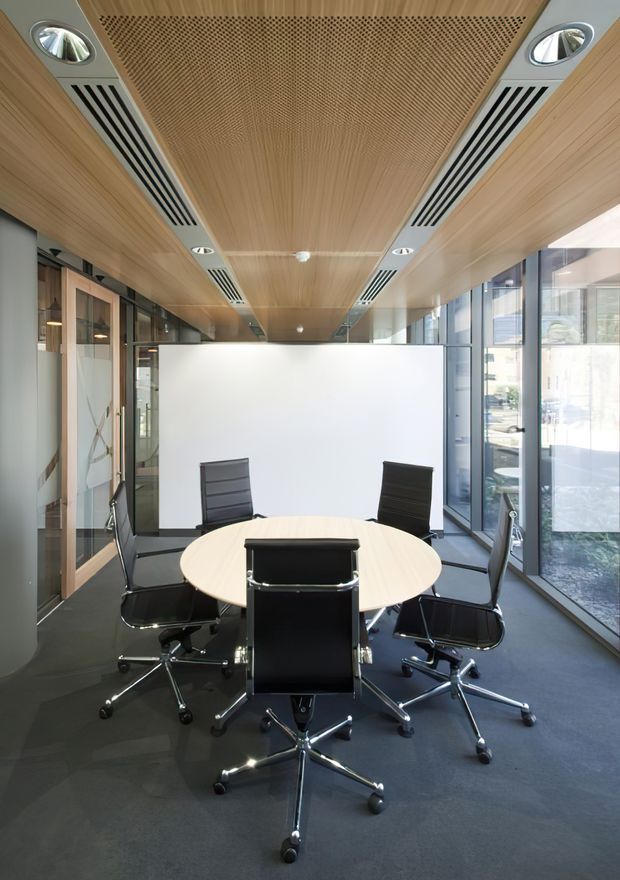A bright contemporary meeting room with a round light-wood conference table and several black office chairs. The room features floor-to-ceiling glazing, a dark carpeted floor, extensive light timber ceiling lining, integrated linear air-conditioning vents, recessed downlights, and a large white presentation screen. Glass partitions and office spaces are visible around the perimeter.