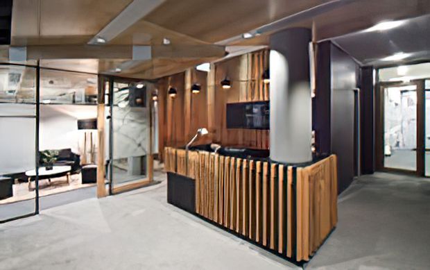 A contemporary commercial reception or concierge area with a sculptural timber-slat front desk, dark charcoal wall panels, exposed warm timber ceiling elements, pendant lights, and glazed partitions. A lounge area is visible to the left, while a corridor and glazed entrance are visible to the right. The space has a refined, modern hospitality or workplace fit-out with strong timber detailing and subdued lighting.