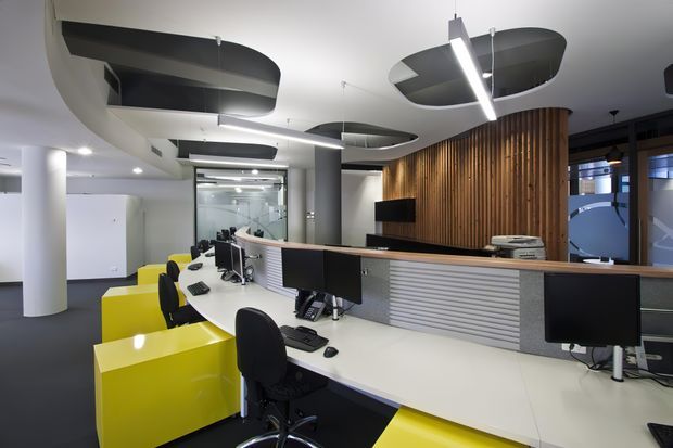 A contemporary commercial office reception or open-plan workspace with a long white workstation and multiple computer monitors, black office chairs, and bright lime-yellow storage or desk modules. The space features white cylindrical columns, a curved slatted timber feature wall, grey ribbed joinery, glass partitions, and suspended linear lighting with sculptural dark acoustic ceiling panels.
