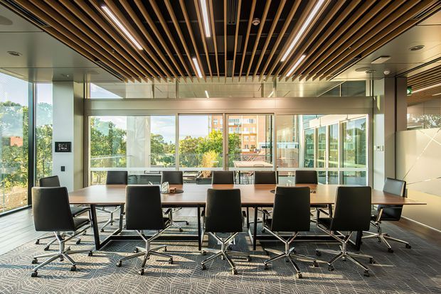 Contemporary commercial conference room with a long rectangular meeting table surrounded by black swivel chairs. The room features floor-to-ceiling glazing, a patterned carpet, pale walls, and a striking ceiling of exposed parallel timber-toned acoustic slats with integrated linear lighting. Greenery and neighboring buildings are visible outside.