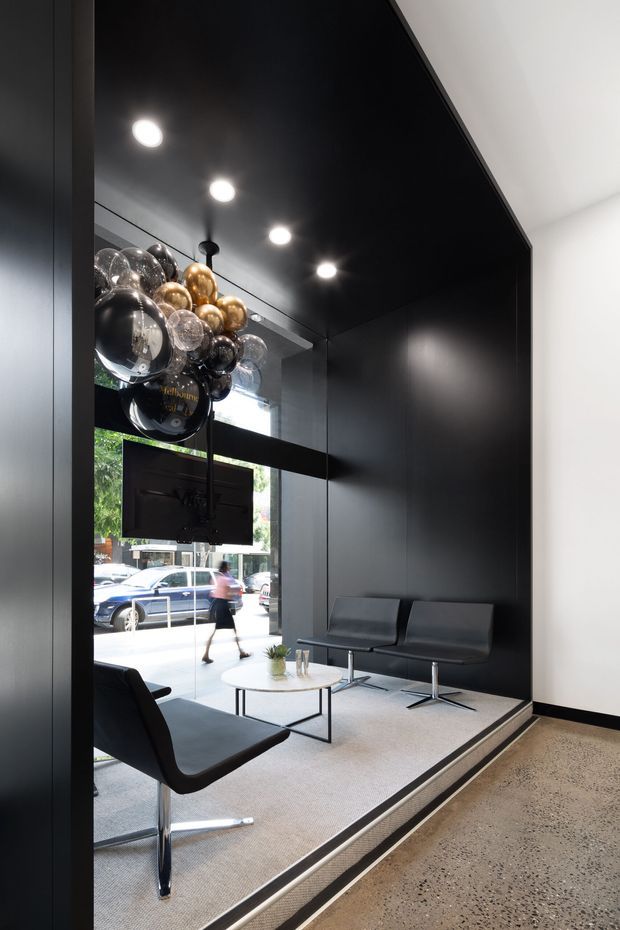 A sleek, compact commercial waiting lounge or reception area with black wall and ceiling panels, large street-facing glazing, contemporary black lounge seating, a small white coffee table, and a dramatic cluster pendant light made of reflective spherical bulbs. The space is raised slightly above a polished concrete circulation floor and has a restrained monochrome palette with strong natural light from the storefront.