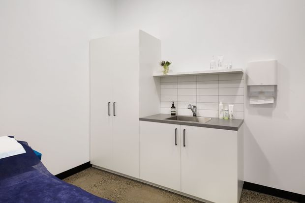Compact, bright clinical-style kitchenette or utility joinery area beside a bed. White full-height cupboards and lower cabinets surround a dark grey benchtop, stainless-steel sink and tap, open shelf with white tiled splashback, wall-mounted paper-towel dispenser, and minimal accessories. The edge of a bed is visible in the foreground, suggesting a hospital, healthcare, or accommodation setting.