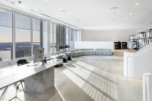 Bright, spacious contemporary open-plan office interior with floor-to-ceiling glazing overlooking a distant landscape. The room features pale timber flooring, white walls and ceiling, minimalist workstations, a large curved sectional sofa, a glass-and-metal stair or mezzanine structure, and strong linear sunlight and shadow patterns across the floor.