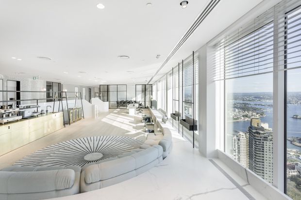 Bright, expansive high-rise interior with floor-to-ceiling glazing, panoramic city and waterfront views, a large curved modular sofa, and an open kitchen or bar area. The predominantly white space features abundant daylight, linear window blinds, polished light flooring, and a dramatic circular radial-pattern rug.