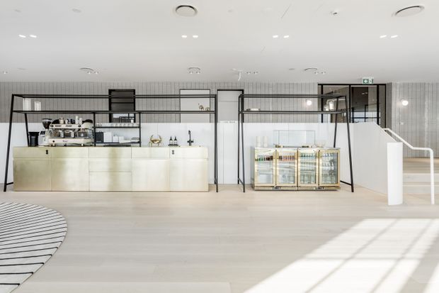 Bright, open-plan contemporary café or hospitality space with a pale timber-toned floor, white counter, black-framed glazed partitions, open metal shelving, commercial coffee equipment, refrigerated display cabinetry, and a staircase at right. The space has a clean, refined fit-out with abundant recessed ceiling lighting and strong natural light.