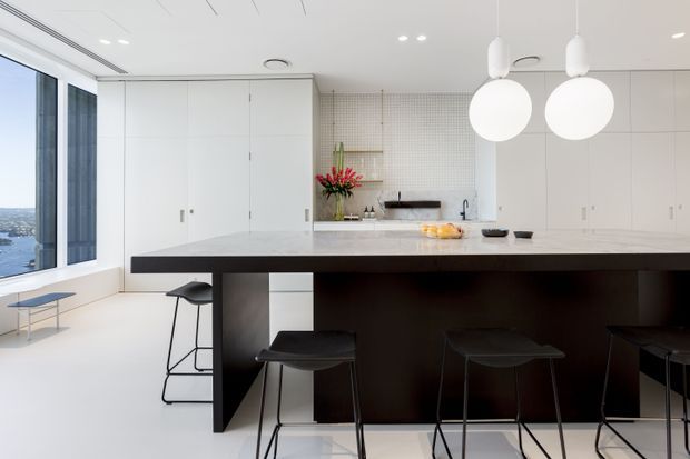 A bright, minimalist contemporary kitchen with extensive white floor-to-ceiling cabinetry, a large dark island with integrated seating, pale stone benchtops, globe pendant lights, and a tiled splashback. Floor-to-ceiling glazing at left provides a coastal or waterside outlook.