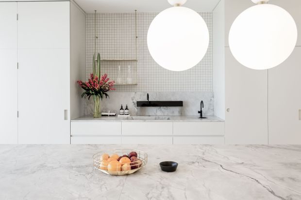 Bright contemporary kitchen with extensive white cabinetry and a large pale marble island benchtop. The rear wall features a marble splashback, white grid-pattern tilework, open shelving, a black wall-mounted tap and basin, and two oversized spherical pendant lights. A fruit bowl and small bowl sit on the island, while a tall floral arrangement adds colour.