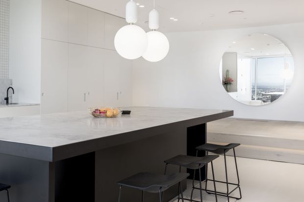 Bright minimalist kitchen with a large pale stone island, dark integrated base, black metal bar stools, full-height light cabinetry, white globe pendant lights, and a circular interior window opening to a bright adjoining space. A distant cityscape is visible through the glazing in the reflection.