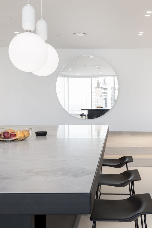 Bright, minimalist contemporary kitchen centered on a large pale stone island with dark cabinetry and three black bar stools. White globe and cylindrical pendant lights hang above, while a large round wall mirror reflects floor-to-ceiling glazing, additional lighting, and part of the kitchen. Pale timber flooring, white walls, and a clean neutral palette create an airy, refined interior.