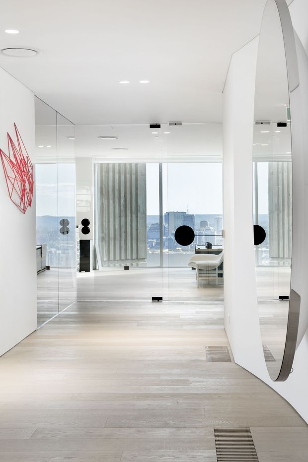 Bright, minimalist contemporary office interior with a pale timber-look floor, white walls and ceiling, full-height glass partitions, and extensive floor-to-ceiling glazing overlooking a city skyline. Mirrored surfaces create reflections through the open-plan space. Abstract red wall artwork provides a small accent, while a desk and seating are visible in the background.