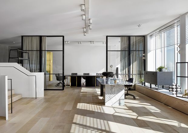 Bright, spacious contemporary open-plan interior with a split-level stair, black-framed glazed partitions, a compact kitchen island, and a home office or study area. Pale timber flooring, white walls, linear track lighting, and extensive louvred glazing create strong geometric shadows across the room.