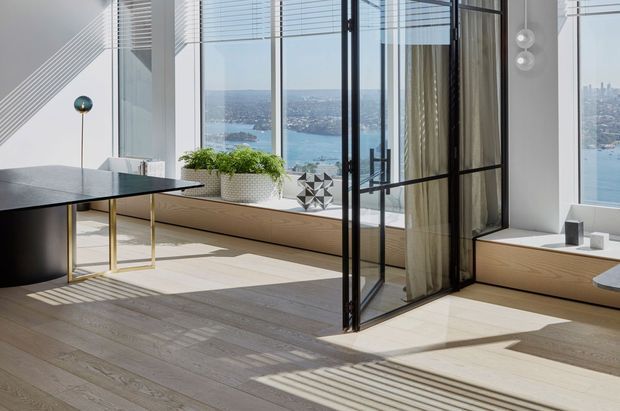 Bright, minimalist contemporary interior with pale timber flooring, expansive floor-to-ceiling glazing, a black-framed glass partition, built-in window seating, and a black dining or work table. The room overlooks a blue harbour and dense urban shoreline, with strong sunlight and linear shadows across the floor.